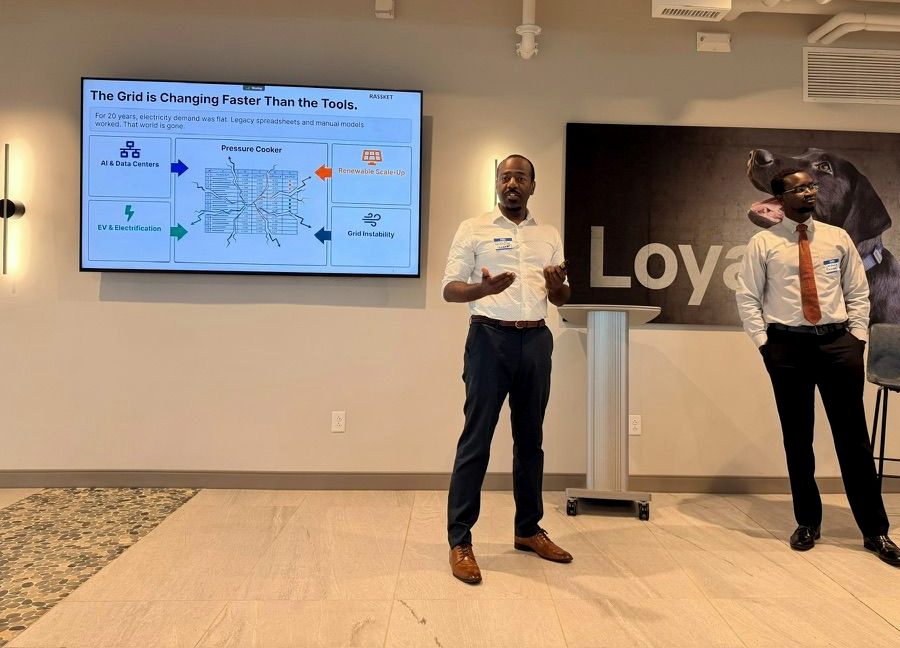 Sankung Fatty presenting on stage in a white shirt, gesturing toward a screen that shows a Rassket slide titled ‘The Grid is Changing Faster Than the Tools’. A colleague in a red tie stands beside him.