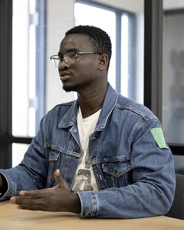 Sankung Fatty in glasses and a denim jacket, seated at a table by a window, mid-explanation with both hands open.